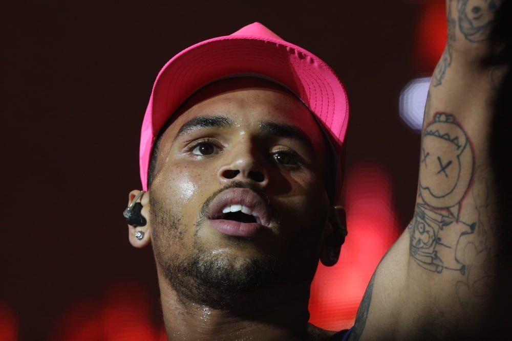 A close-up of Chris Brown performing on stage in a pink cap, one arm raised to reveal tattoos, under red stage lighting.