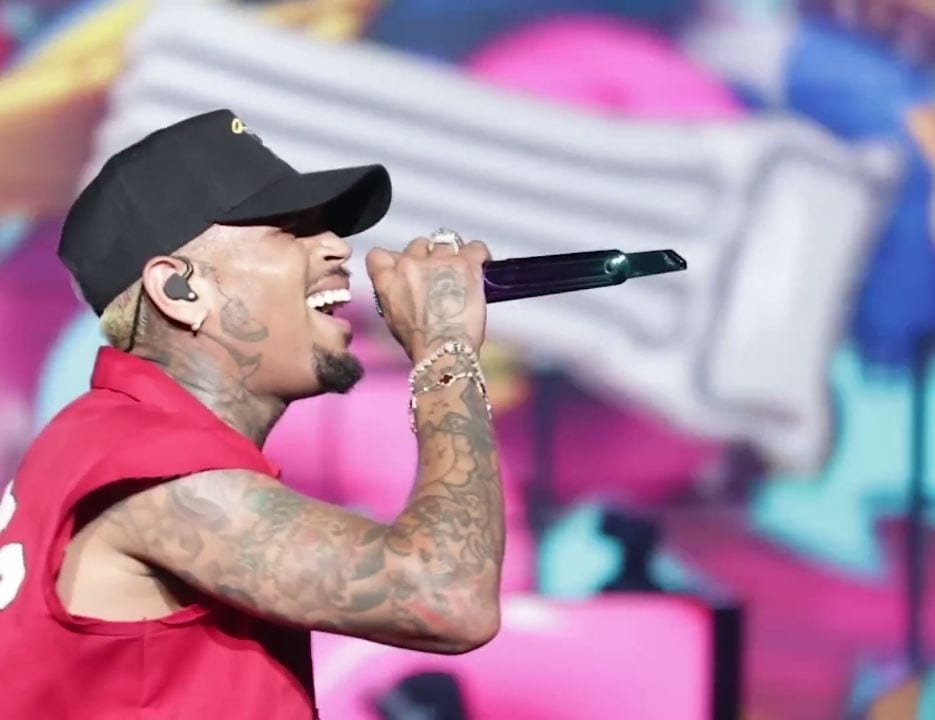 Chris Brown, in a black cap and red tank top, sings into a microphone, his tattooed arm raised.