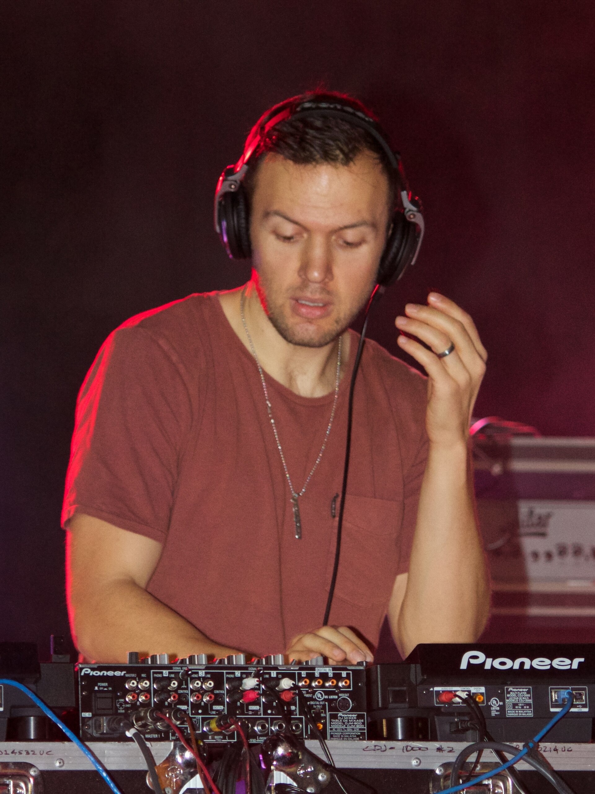 Chris Lake looks down at a Pioneer mixer with headphones on and one hand raised to an earcup, in a plain red t-shirt under red light.