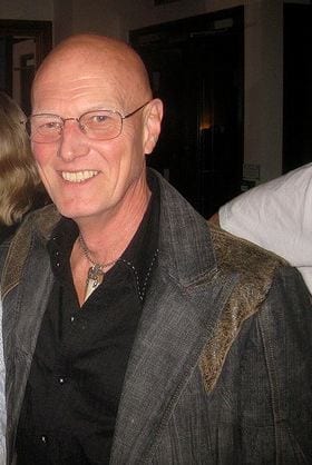 Chris Slade smiles in glasses and a grey fur-trimmed jacket over a black shirt.
