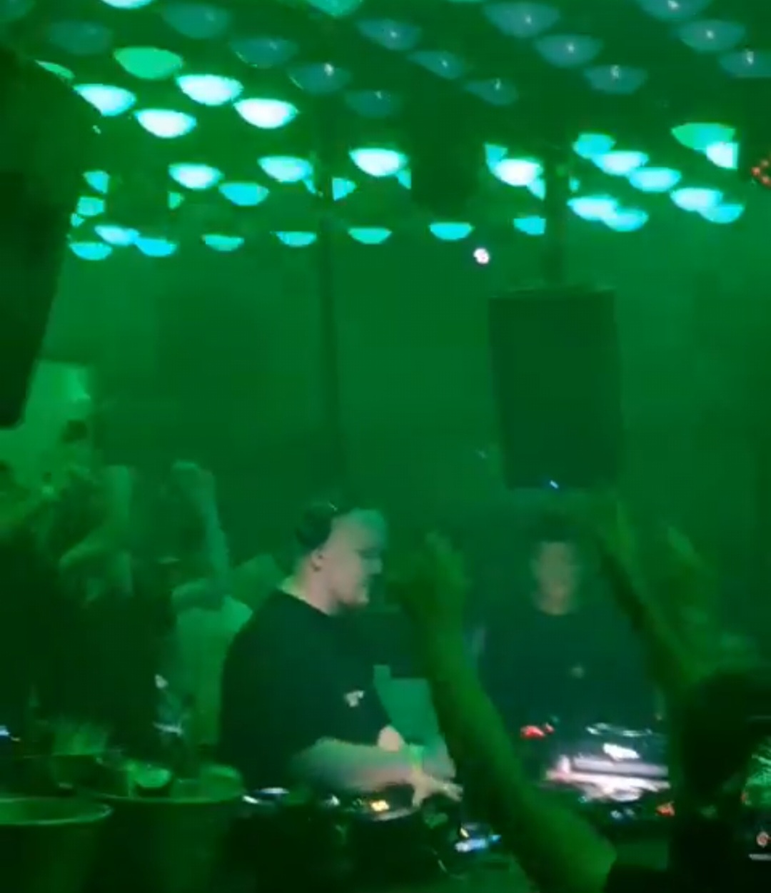 Two figures work a DJ booth in a club lit almost entirely green, the decks and a raised hand picked out against the haze.