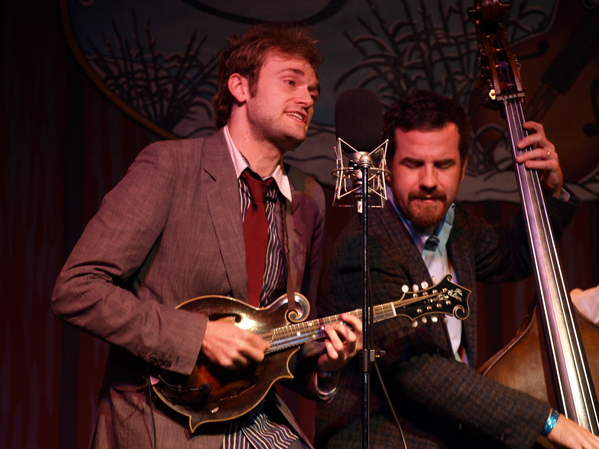 Chris Thile plays mandolin in a grey suit and red tie, singing towards a condenser microphone, with bassist Greg Garrison leaning in behind the same mic.