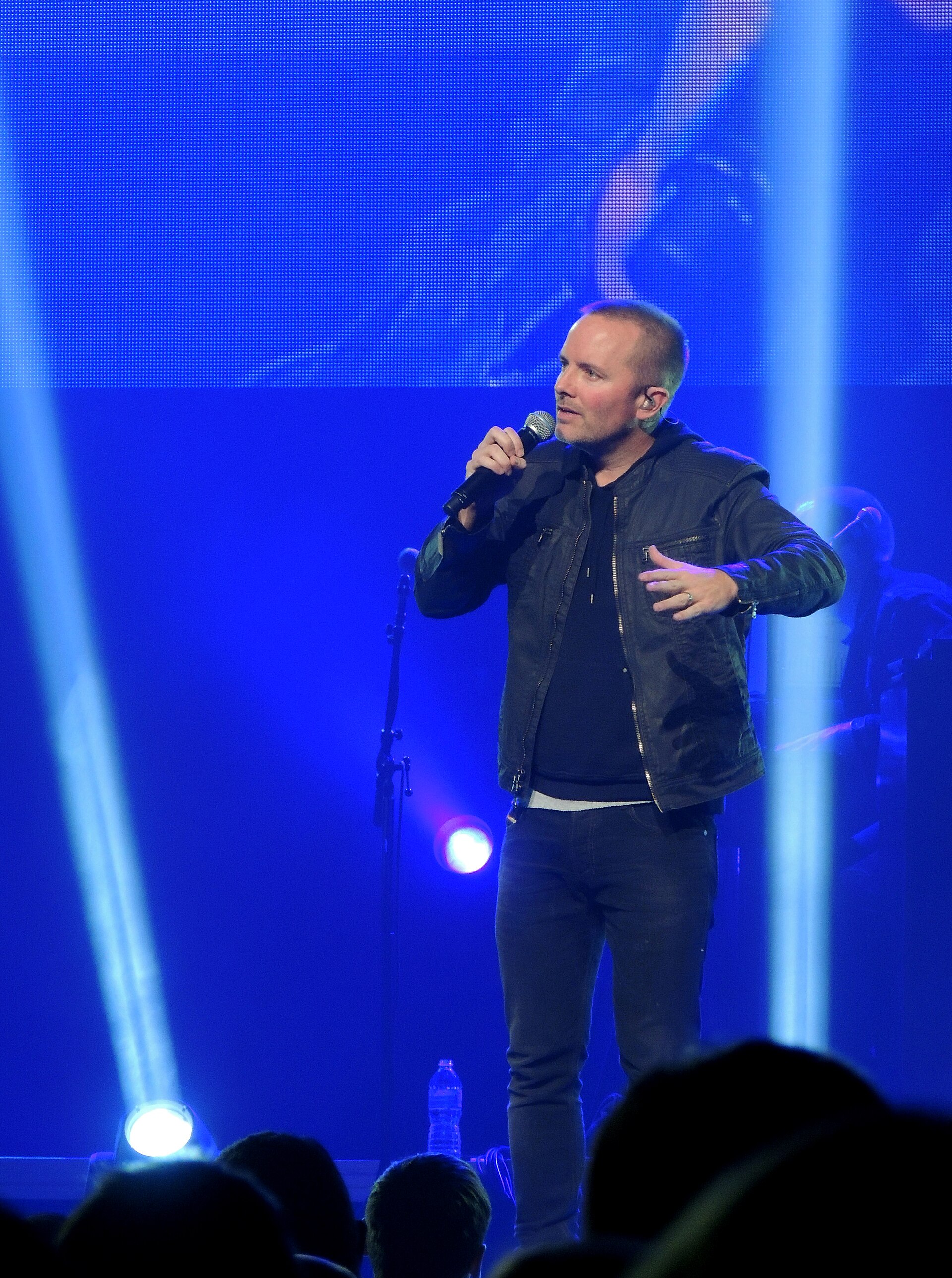 Chris Tomlin sings into a handheld microphone on a stage washed in deep blue light, in a black jacket and jeans with a video screen glowing behind him.