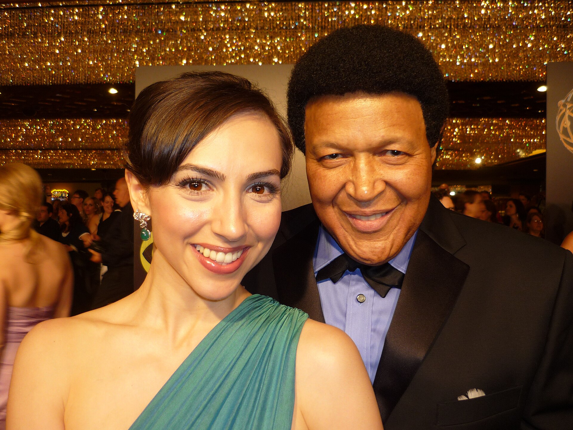 Chubby Checker, in a black tuxedo and bow tie, smiles beside the actress Eden Riegel in a green one-shoulder gown, under gold beaded awards-show decor.