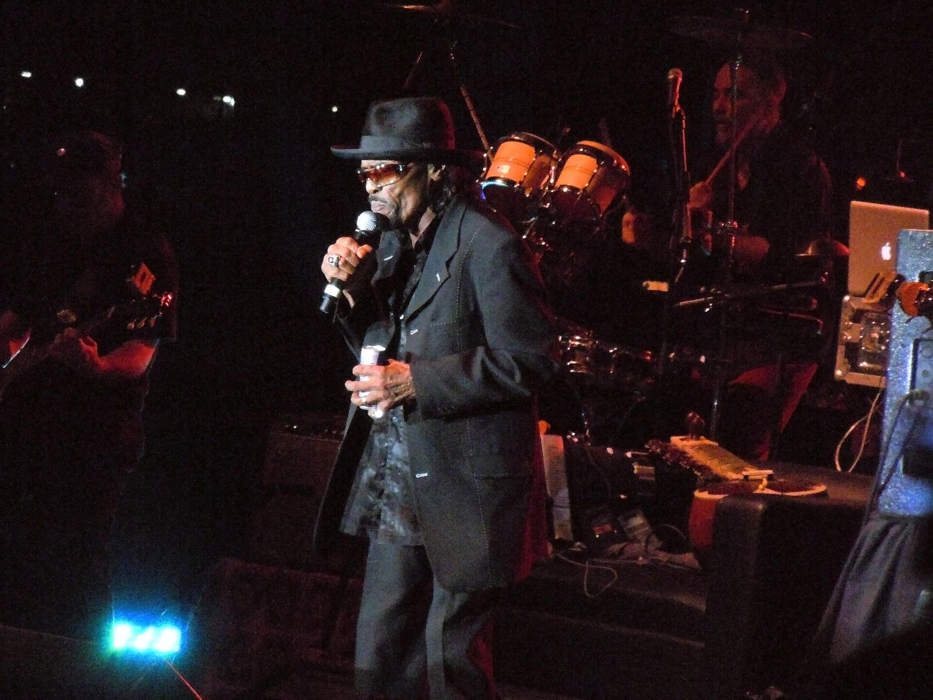 Chuck Brown sings into a handheld microphone in a wide-brimmed hat and tinted glasses on a dark stage, congas and a guitarist behind him.
