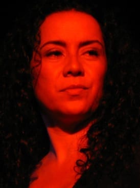 Claudia Acuna, with curly dark hair, is lit entirely in deep red light in a close-up portrait.
