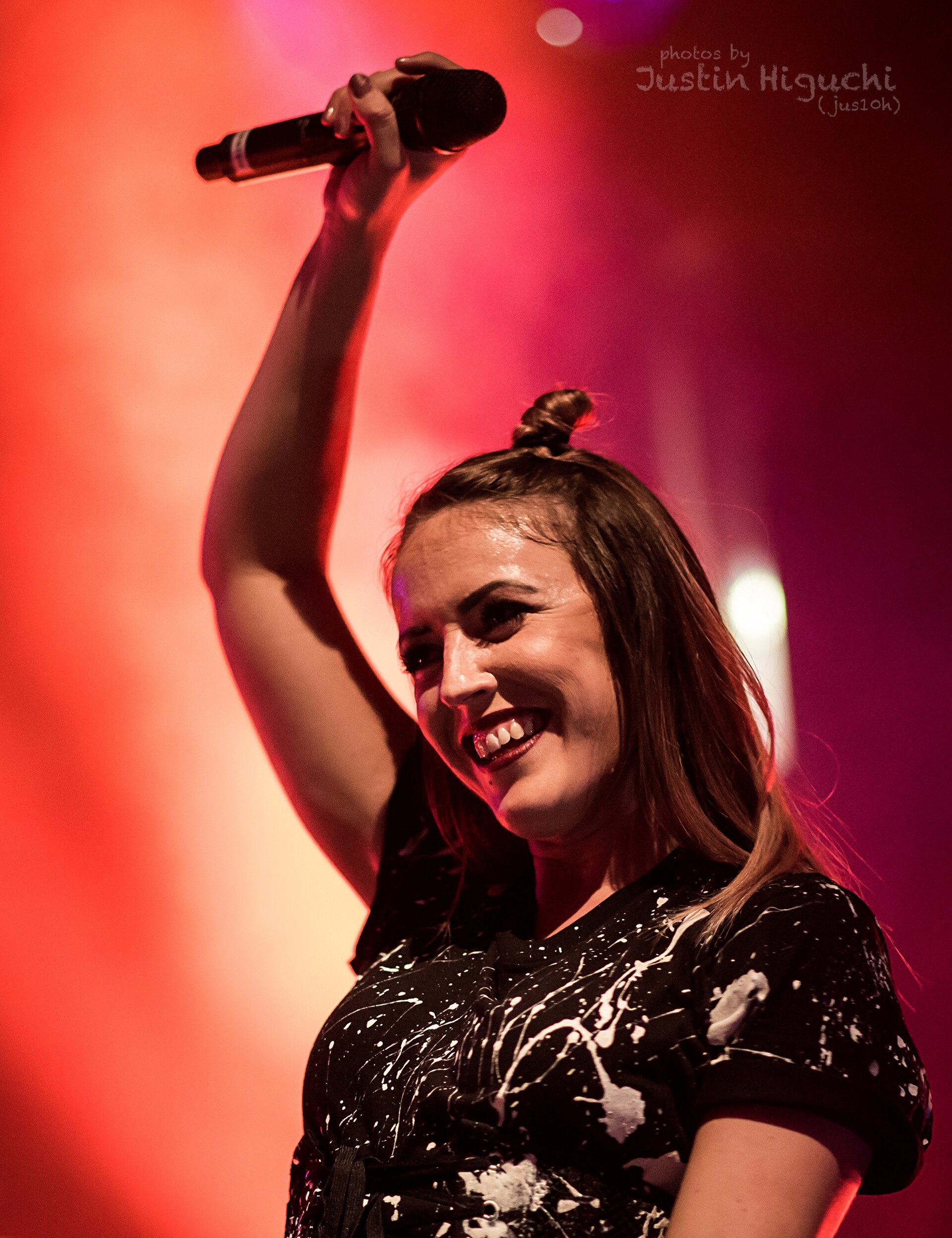 A Clean Bandit singer grins and holds a microphone high overhead in a paint-splattered black top, lit red from behind.