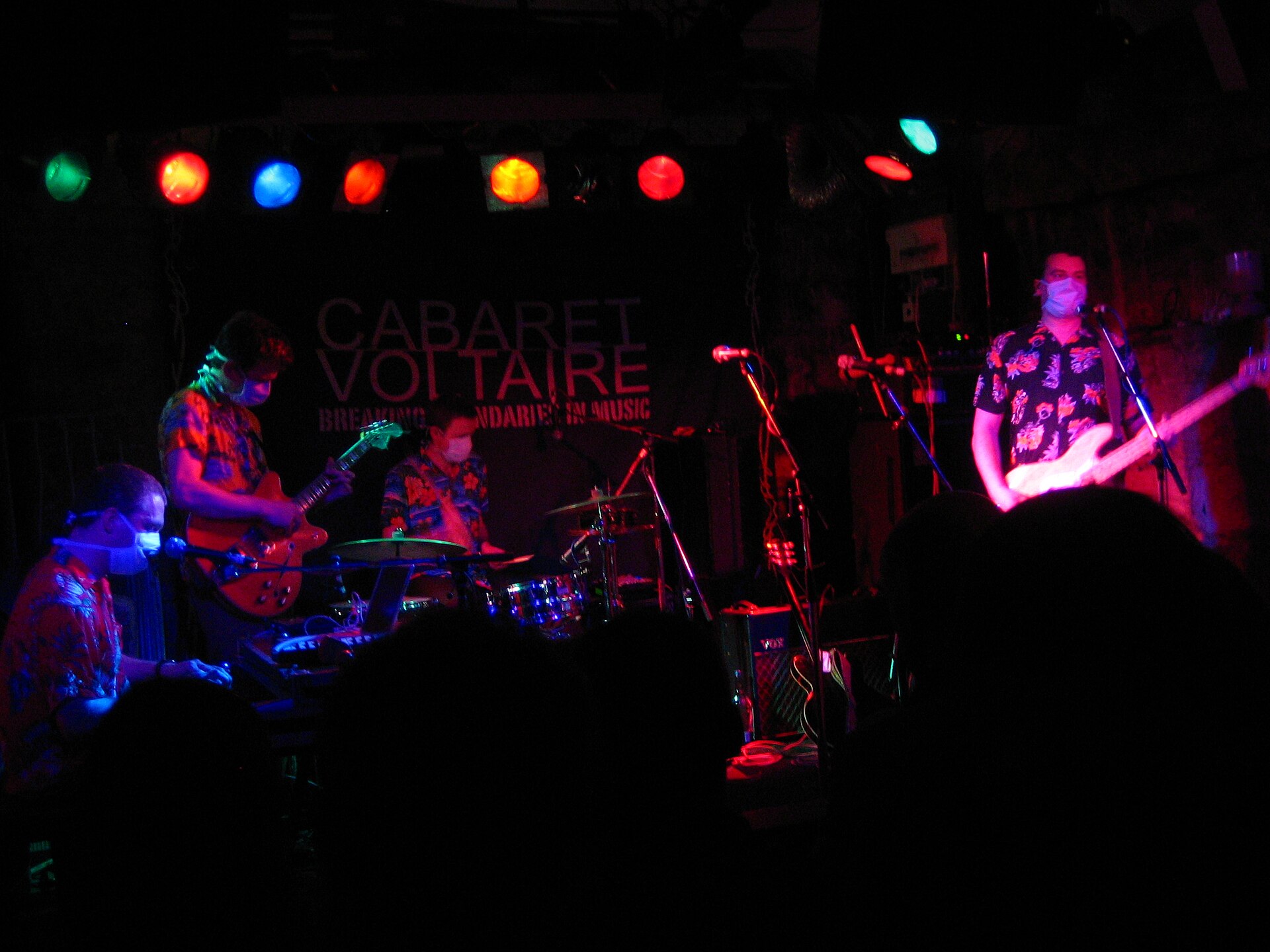 The four members of Clinic play a small club stage in matching floral shirts and surgical masks under a row of coloured lamps.