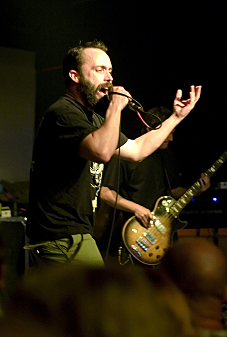 Neil Fallon of Clutch shouts into a handheld microphone with his free hand raised, a guitarist with a sunburst Les Paul behind him and the crowd blurred below.