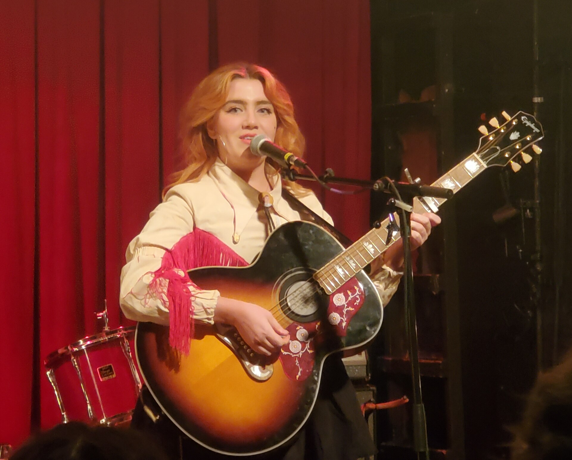 CMAT sings at a microphone in a cream fringed western shirt, playing a sunburst acoustic guitar in front of a red curtain.