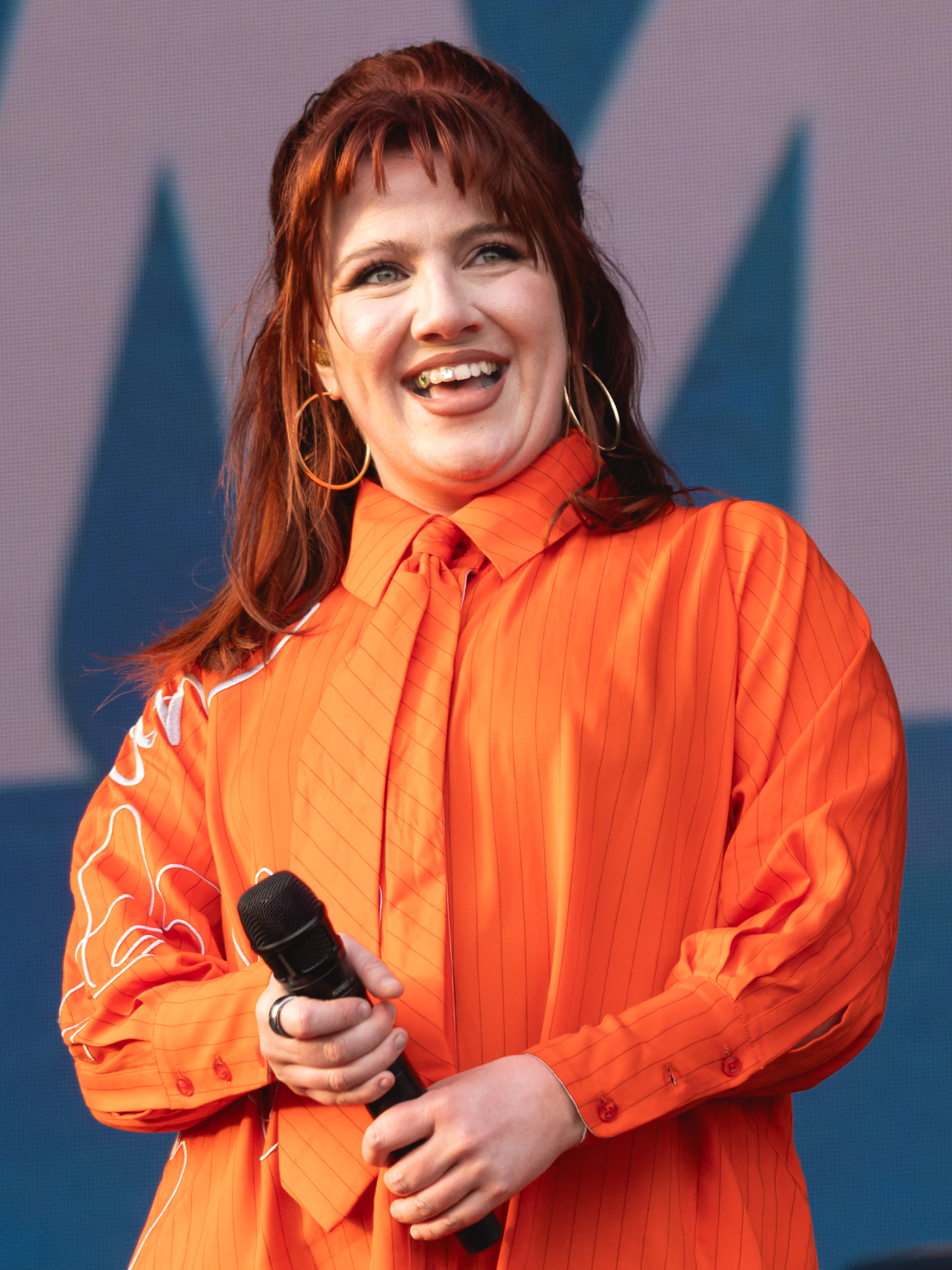 CMAT laughs mid-sentence holding a microphone, with long red hair, large hoop earrings and a bright orange shirt.