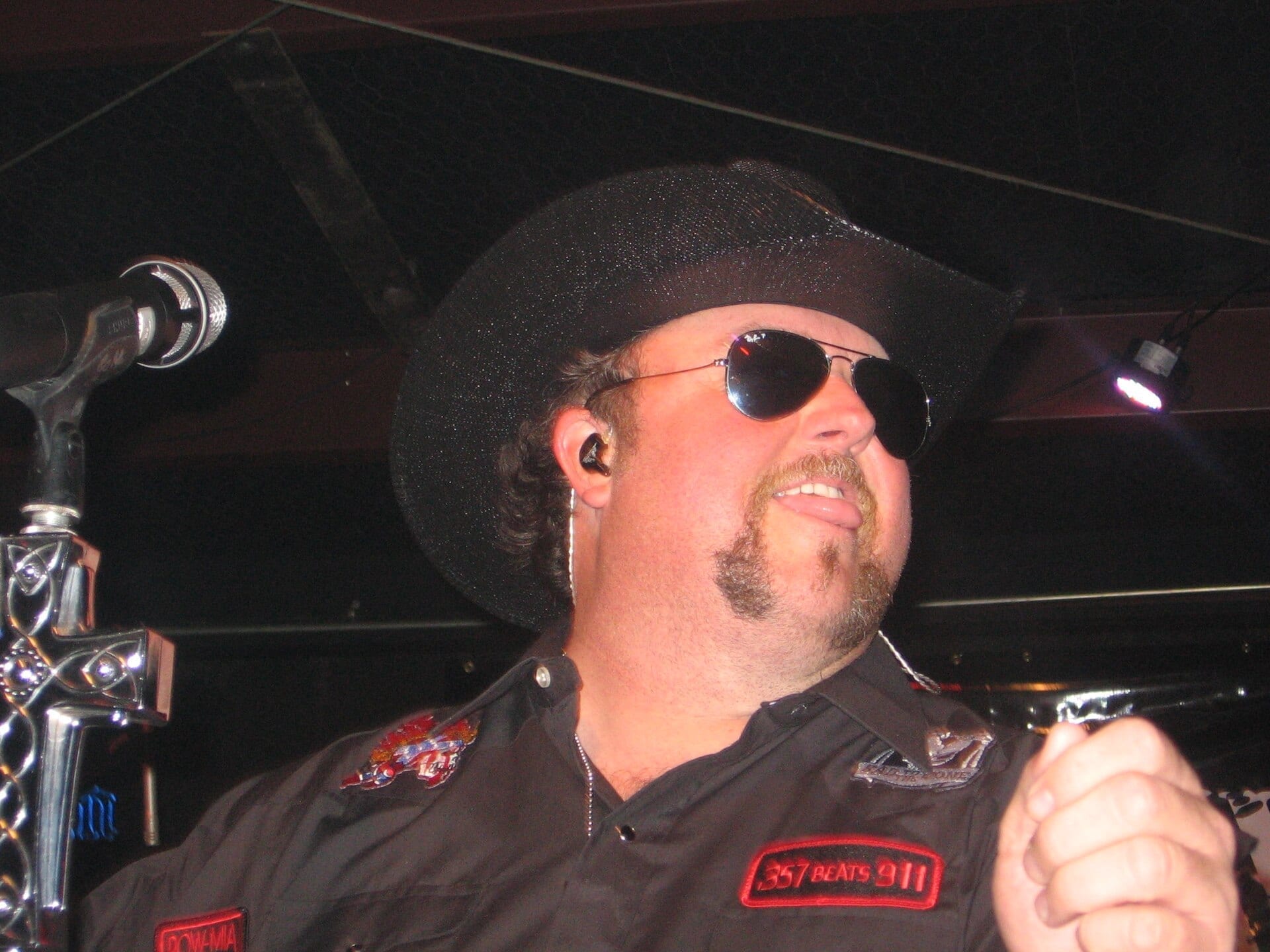 Colt Ford, wearing a black cowboy hat and aviator sunglasses, sings into a microphone on stage in a dark button-up shirt with patches.