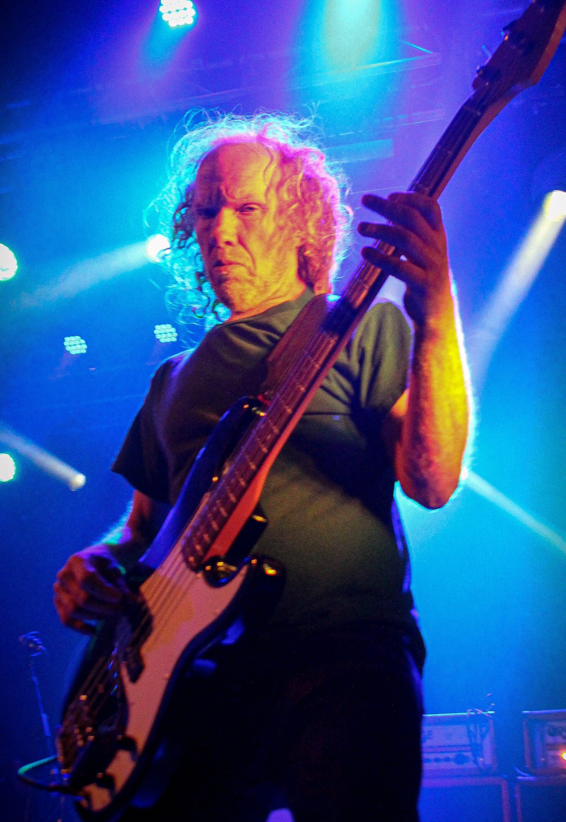 A bassist with long curly grey hair leans into a bass guitar on a club stage, shot from below against blue and magenta lights.