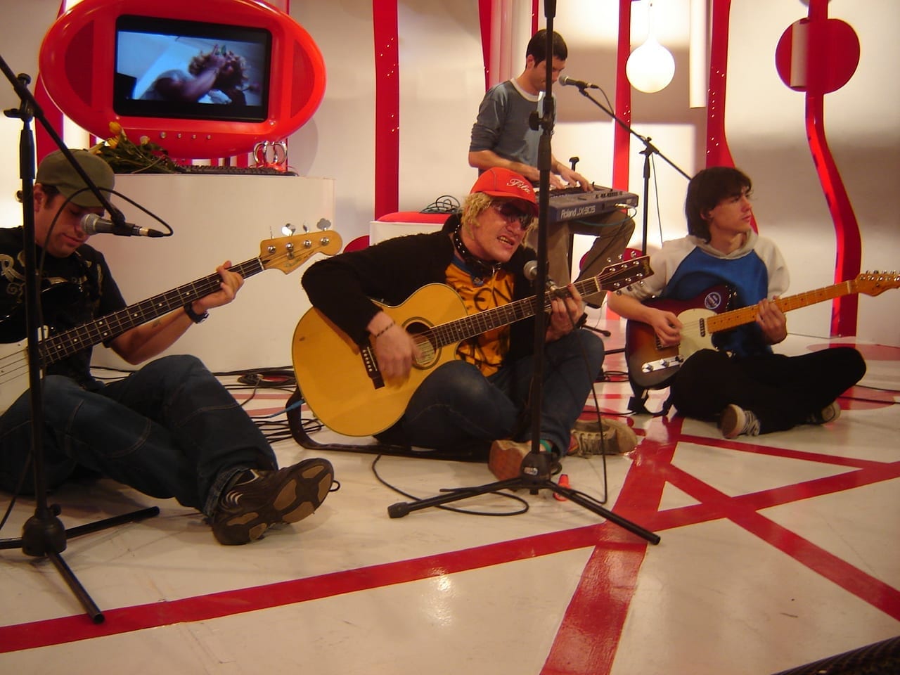 Pity Alvarez sings into a microphone while playing an acoustic guitar, wearing a red cap and sunglasses, backed by his band Intoxicados on a red and white television studio set.