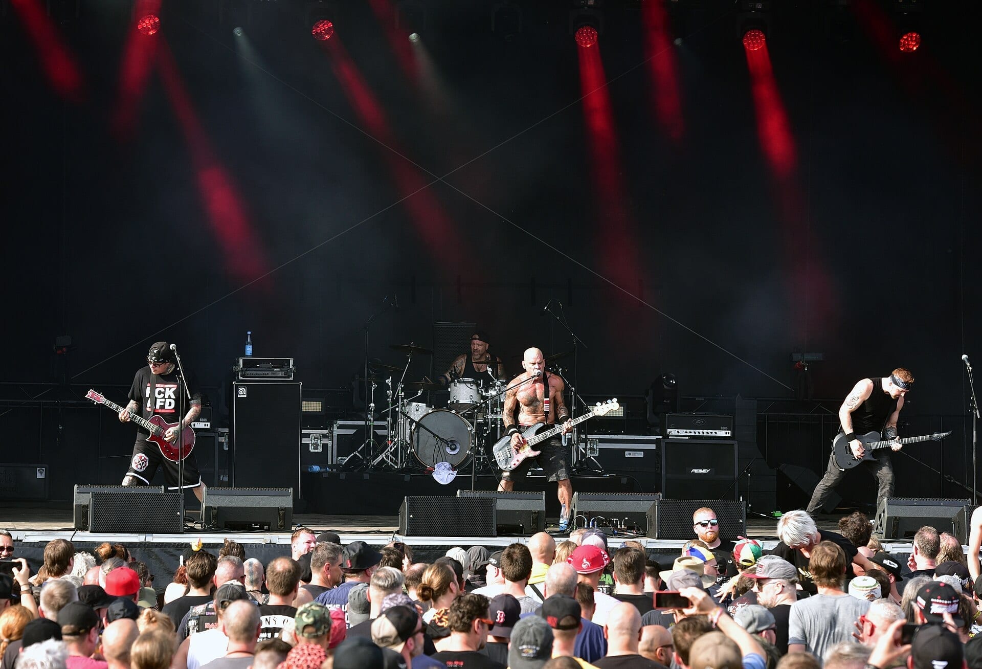 Four members of Cro-Mags perform on an outdoor festival stage in daylight, guitarists flanking a shirtless bassist and vocalist at center, with a large crowd in front of the stage.