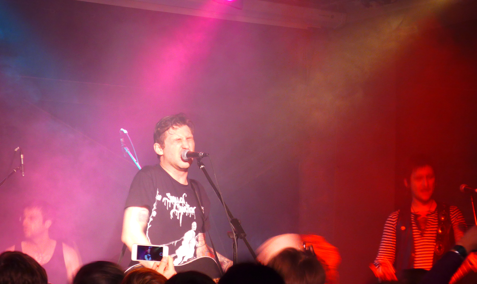 Cult of Youth's singer leans into a microphone with an acoustic guitar, in a black band T-shirt under pink and red stage haze, a bandmate in stripes to the right.