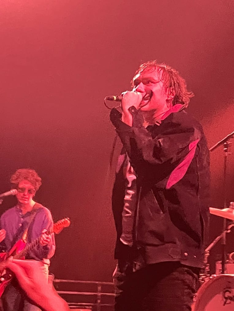 A man with curly hair sings into a microphone under red stage lighting, with a guitarist playing beside him.