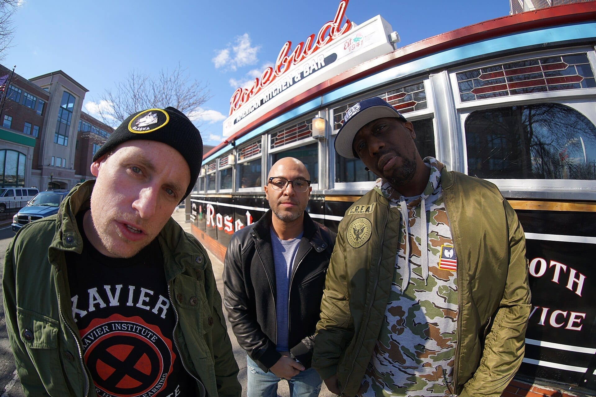 Three members of Czarface stand shoulder to shoulder on a sunny street outside the Rosebud diner, one in a black beanie and Xavier Institute t-shirt, one in a leather jacket, and one in a camouflage jacket with a Wu-Tang patch and a cap reading C.R.E.A.M.