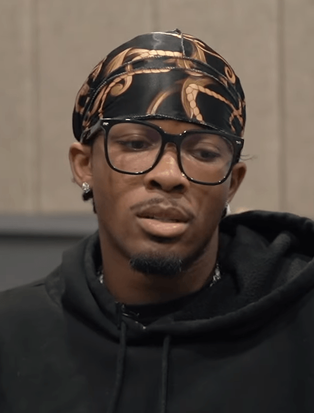 D4vd, wearing a patterned durag, black-framed glasses and a black hoodie, sits for an interview with his hand near his chin.