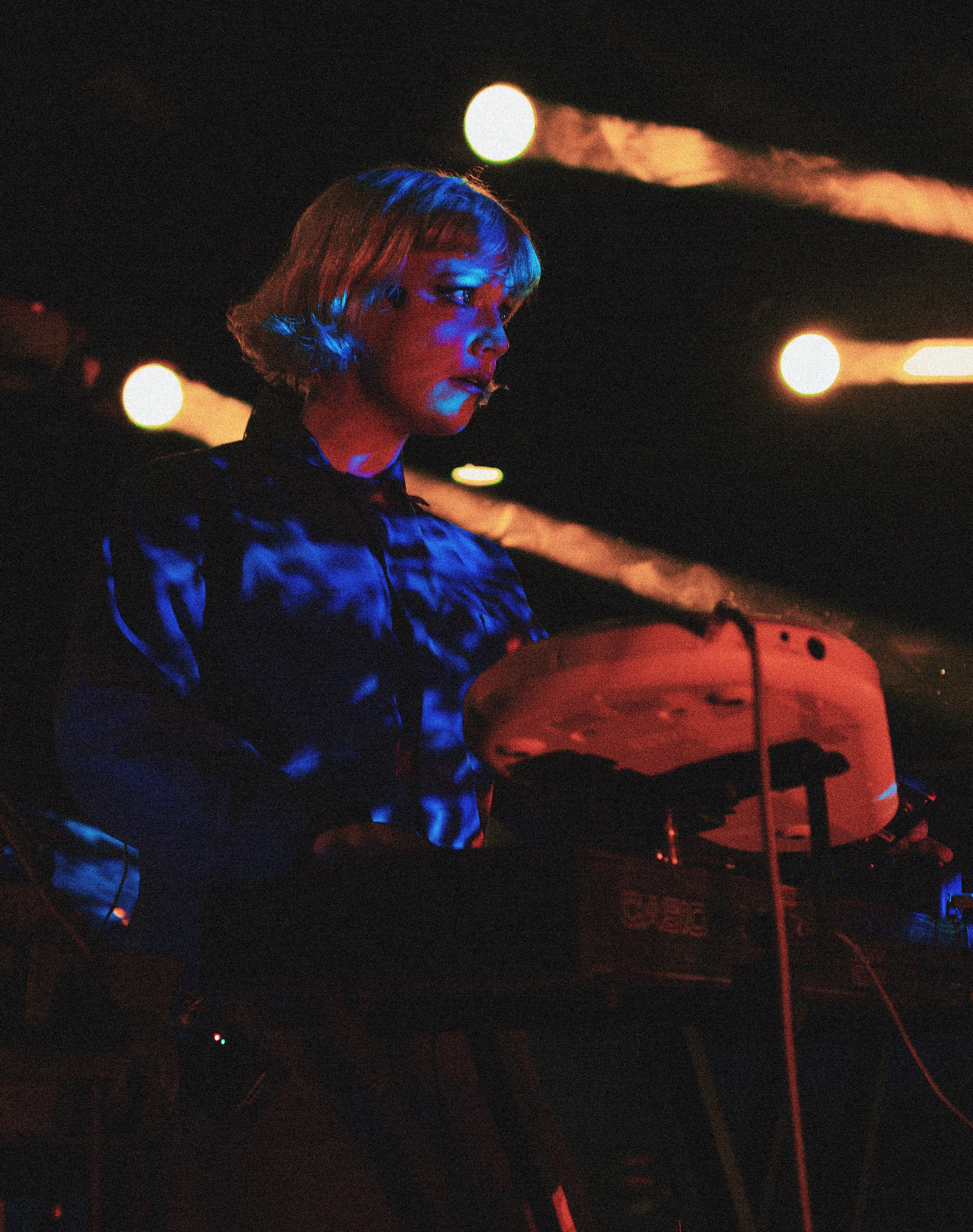 Danz CM stands behind a bank of synthesizers in a headset microphone and a patterned shirt, lit blue against warm stage lamps.