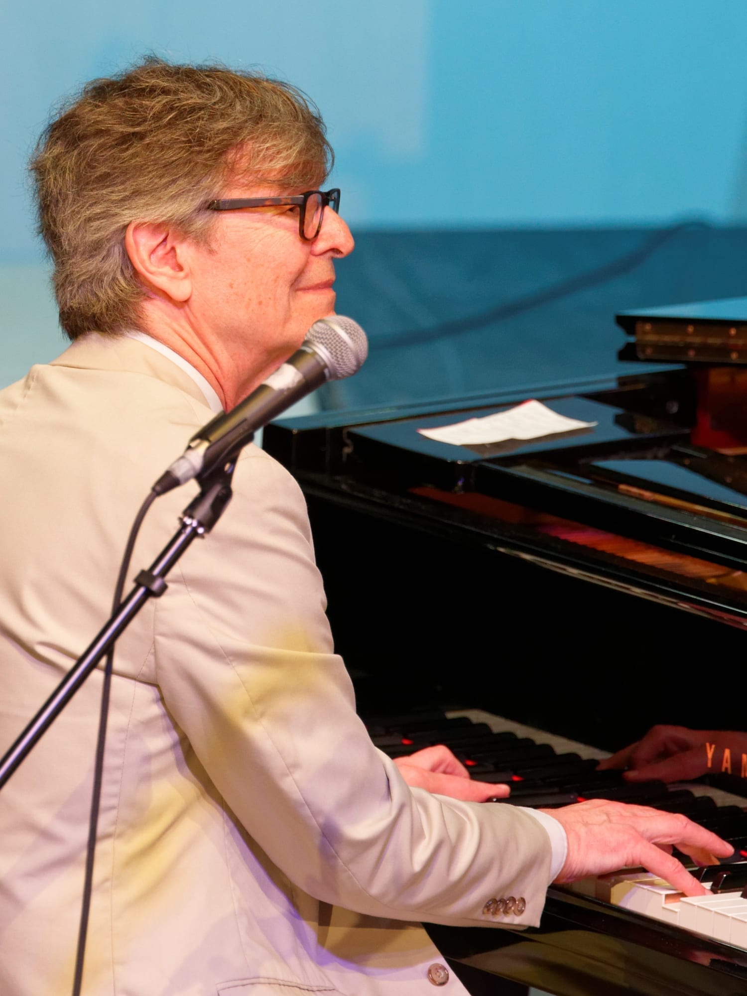 Darius Brubeck plays a grand piano in profile, wearing glasses and a cream blazer, with a microphone positioned in front of him.