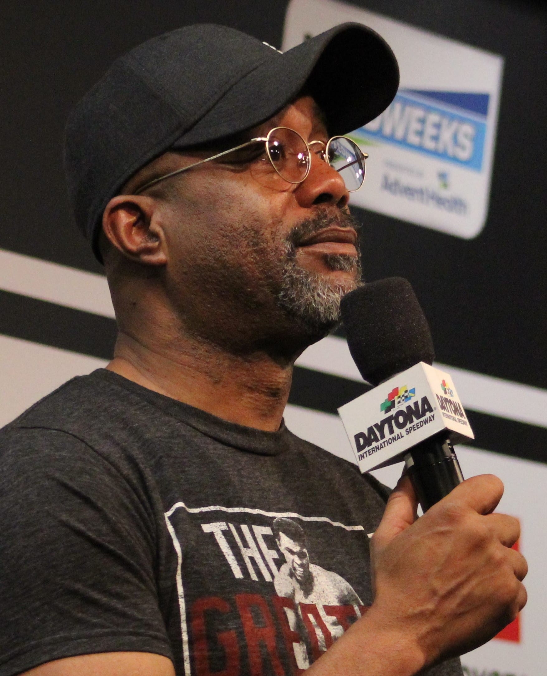 Darius Rucker, wearing round glasses and a cap, speaks into a Daytona International Speedway branded microphone.