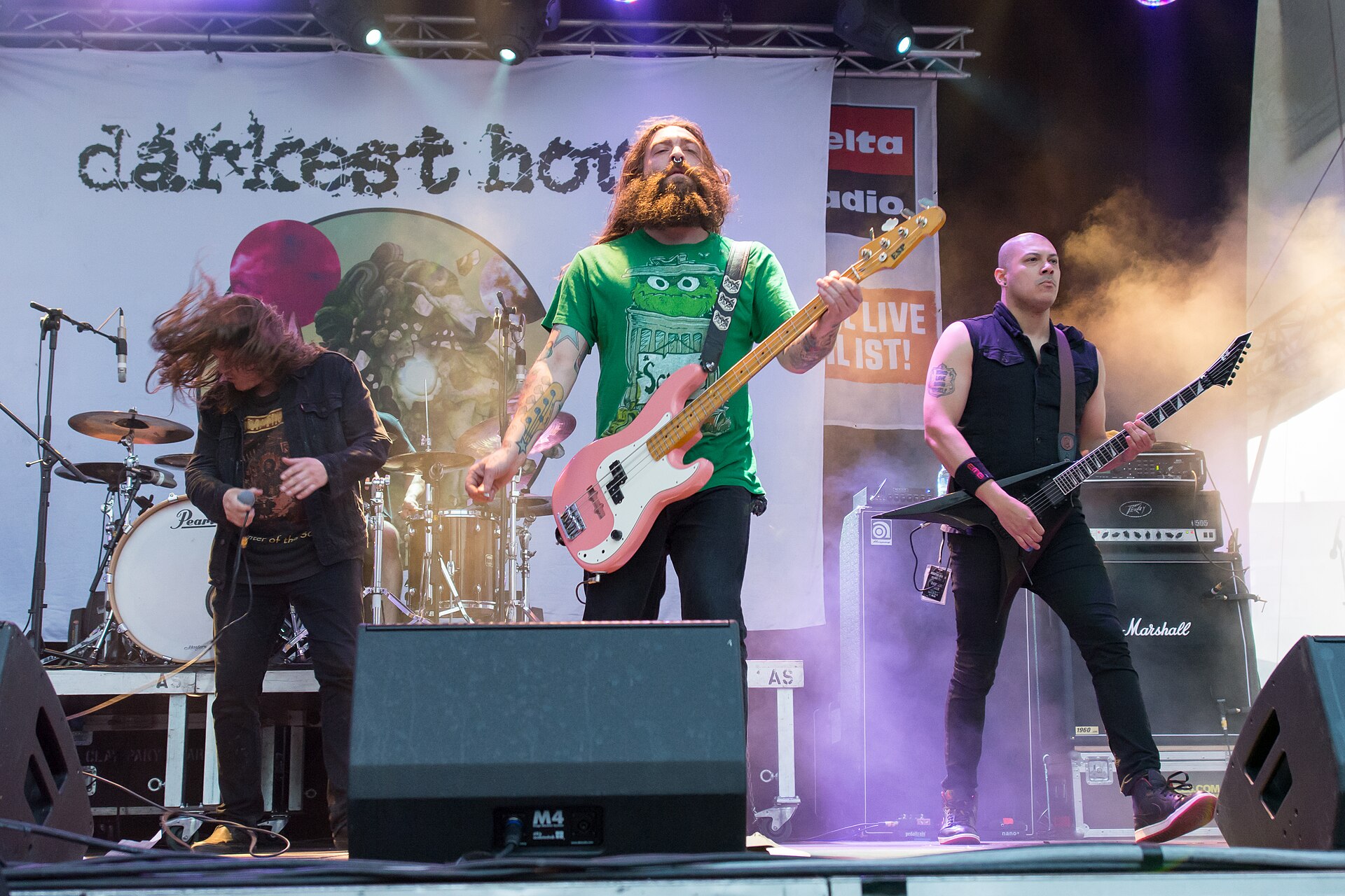 Darkest Hour play a festival stage in front of a banner bearing the band's name, the bassist in a green T-shirt with a pink bass, a guitarist in black to his right.