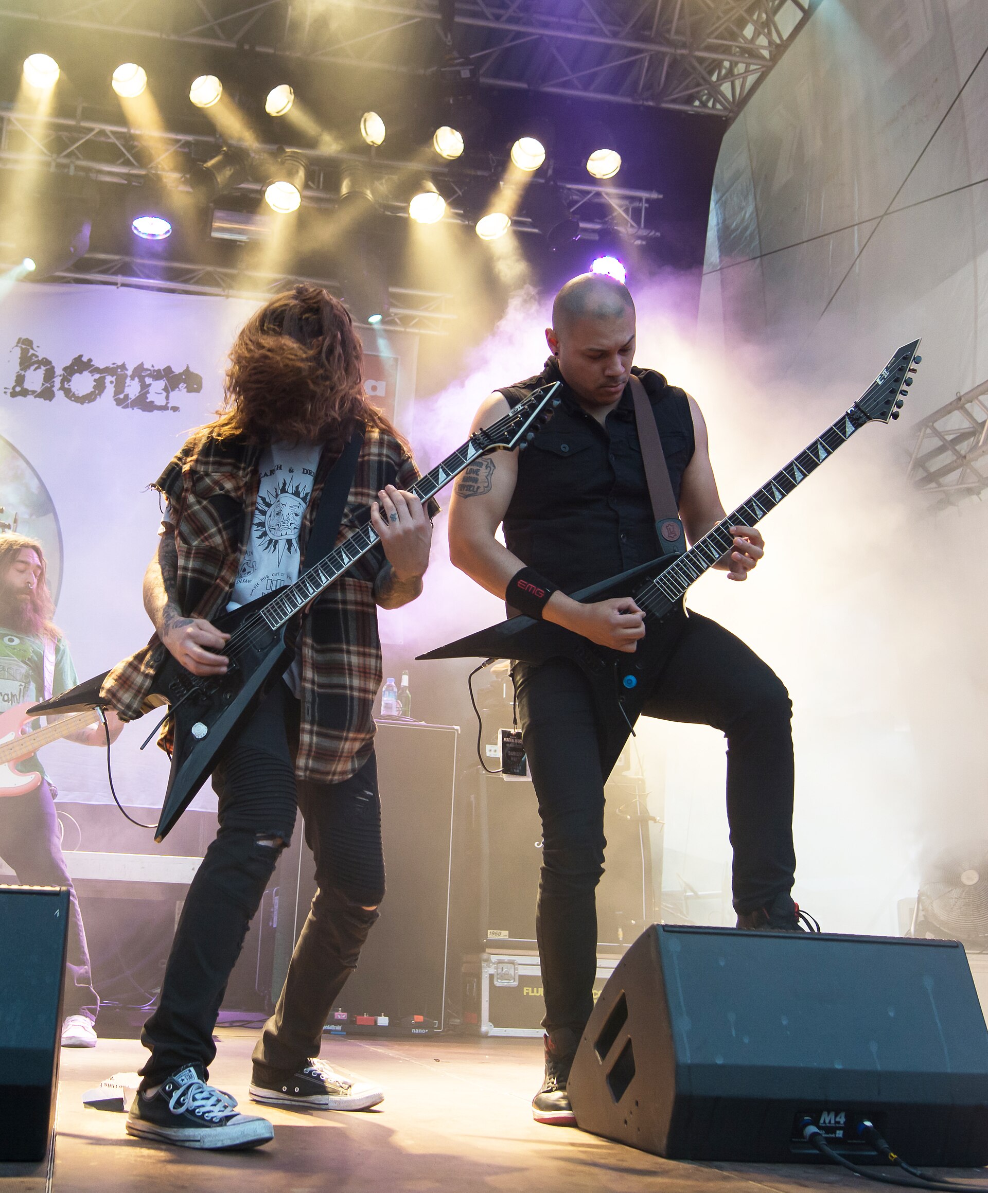 Two Darkest Hour guitarists play black V-shaped guitars head-down in stage smoke, one in a plaid shirt with hair over his face, the other in a cut-off black shirt.