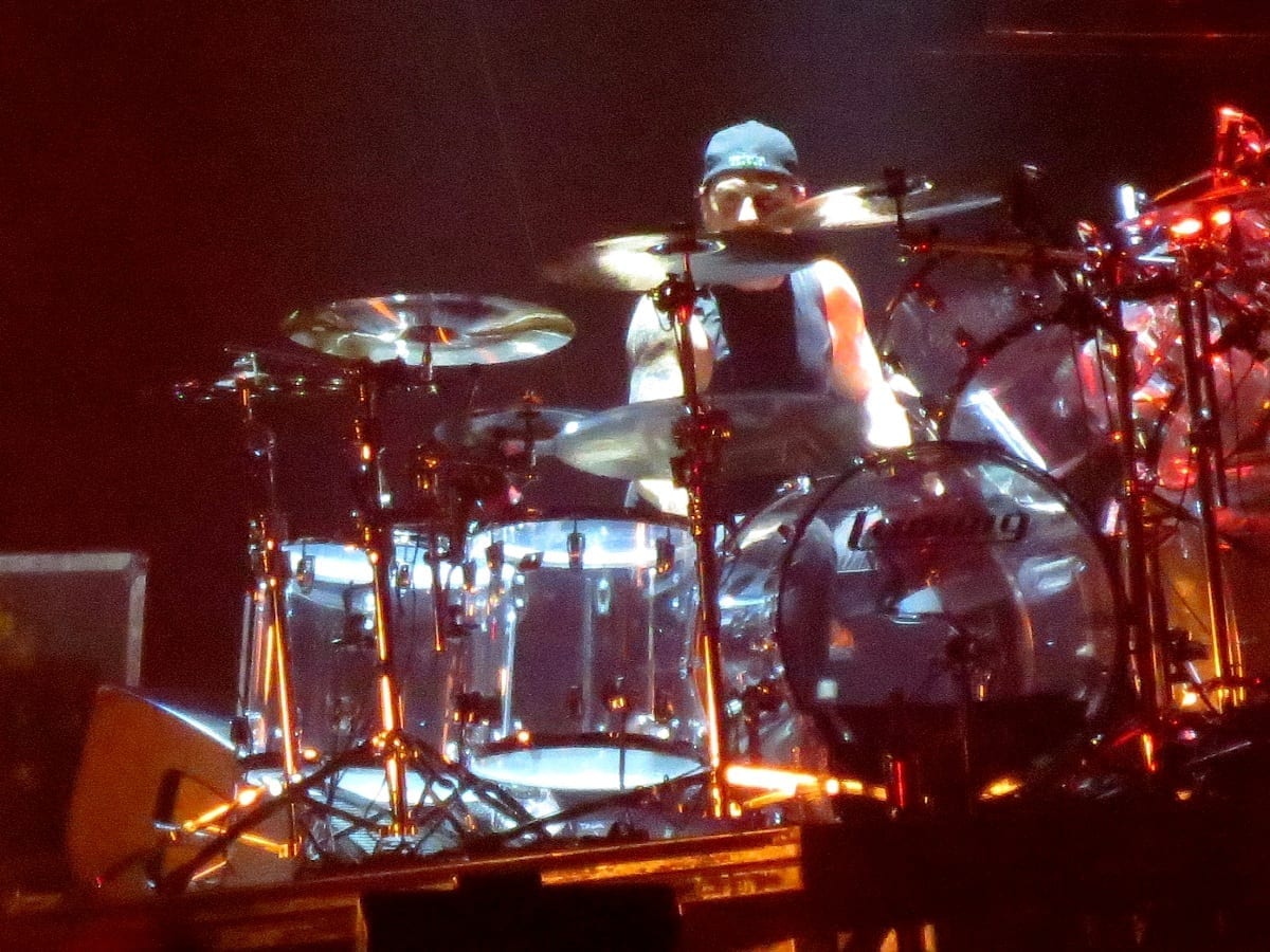 Dave Lombardo plays drums on a chrome-finished kit, drumsticks blurring in motion above the cymbals, lit by red and white stage lights.