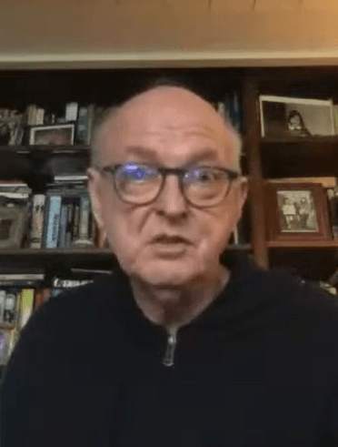 Dave Marsh, wearing glasses and a dark half-zip sweater, speaks on a video call in front of a bookshelf lined with books and framed photos.