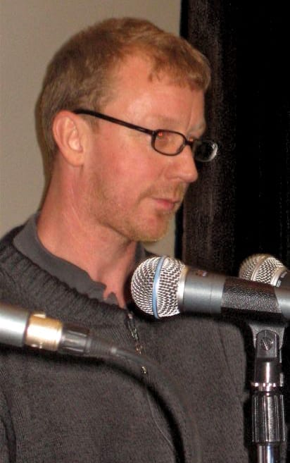 Dave Rowntree, wearing glasses and a gray sweater, speaks close to a microphone on a stand.