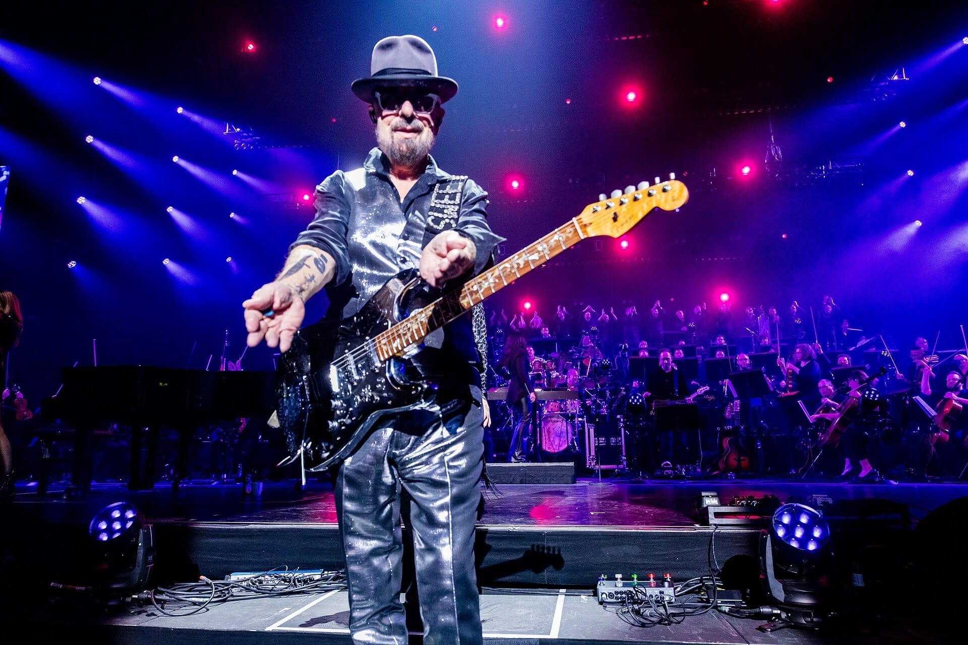 Dave Stewart, in a fedora, sunglasses and metallic silver suit, plays an electric guitar in front of a full orchestra on stage.
