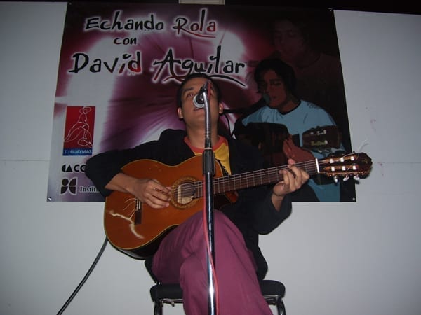 David Aguilar sings into a microphone while playing an acoustic guitar, seated in front of a banner reading 'Echando Rola con David Aguilar'.