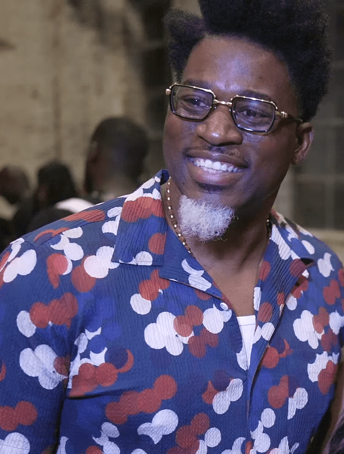 David Banner smiles off-camera in gold-rimmed rectangular glasses and a blue shirt patterned with red and white circles, a white goatee under his chin.