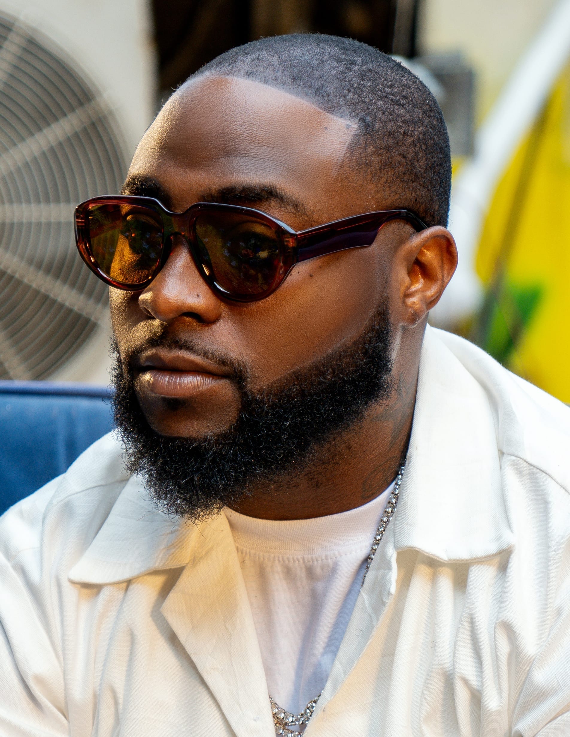 Davido, wearing tinted sunglasses and a white shirt jacket, looks off to the side in a close-up outdoor portrait.