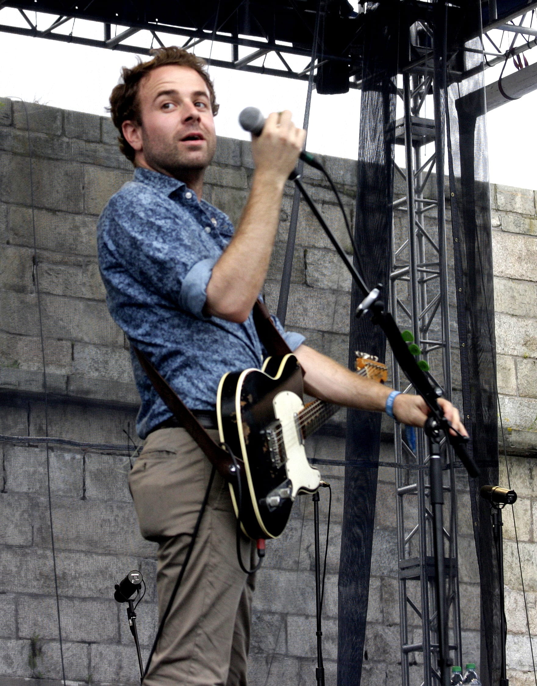 Taylor Goldsmith sings into a handheld microphone with an electric guitar slung over a patterned blue shirt.