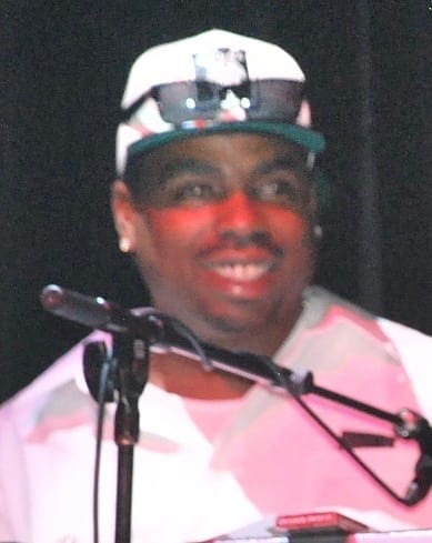 Daz Dillinger smiles into a microphone, wearing a white and green cap with sunglasses perched on the brim, under red stage light.