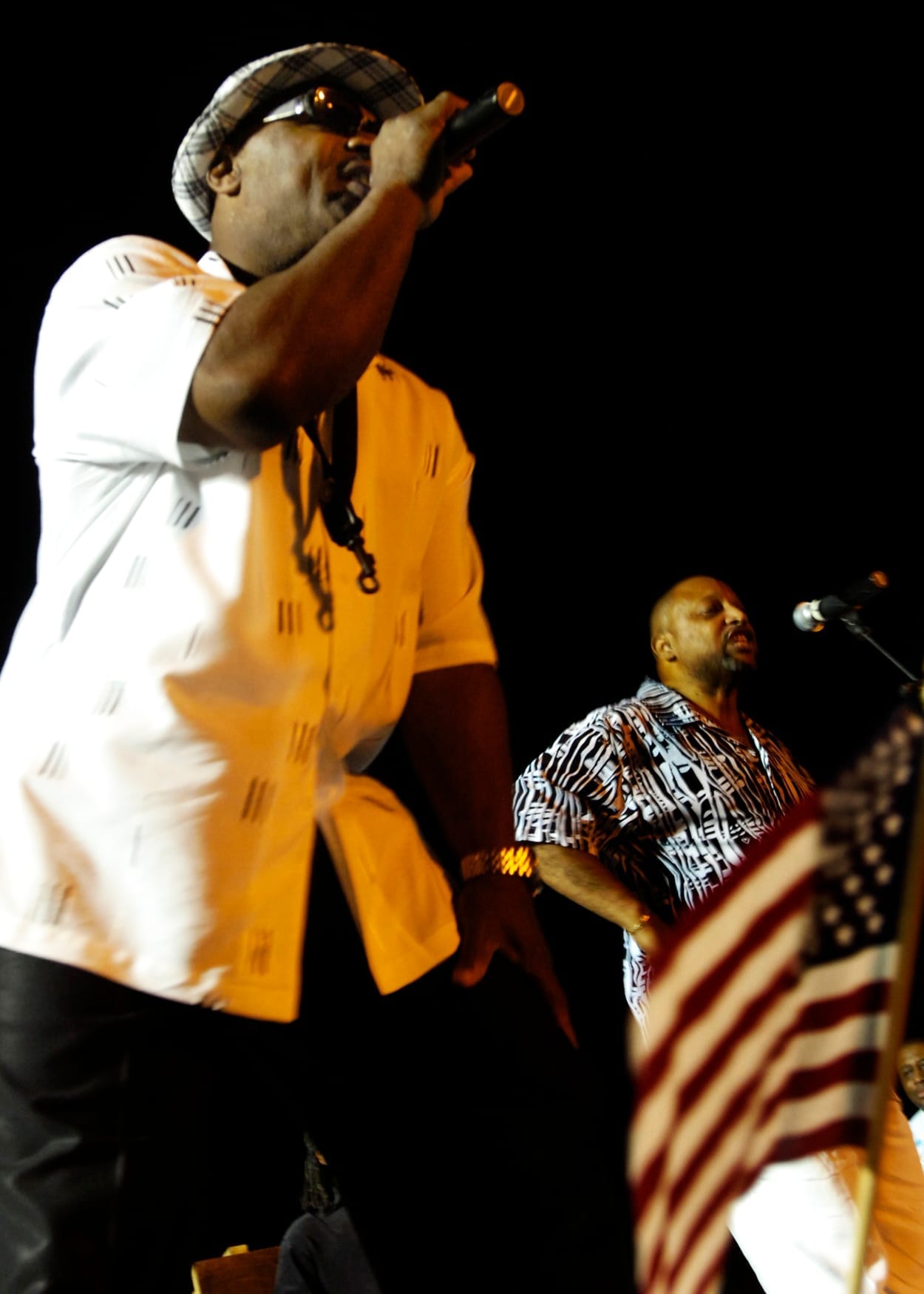 Bobby Harris of the Dazz Band sings into a microphone on an outdoor stage at night, wearing a patterned cap and sunglasses, with a bandmate and an American flag behind him.