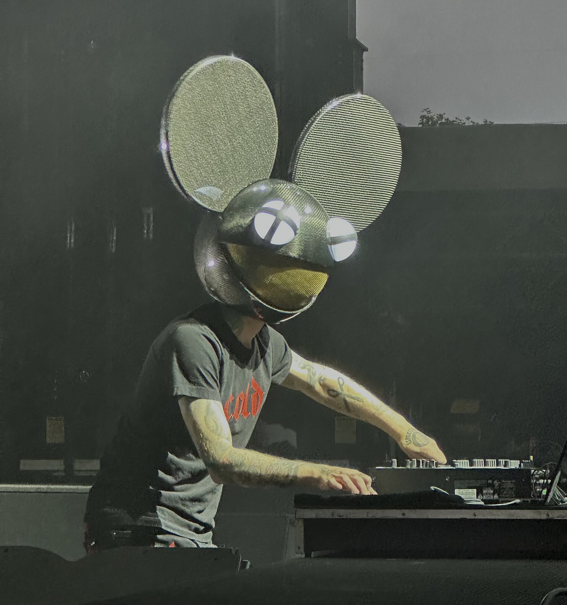 deadmau5 works a DJ mixer in his oversized mouse-head helmet, the eyes and mouth lit up, tattooed arms reaching across the controls.