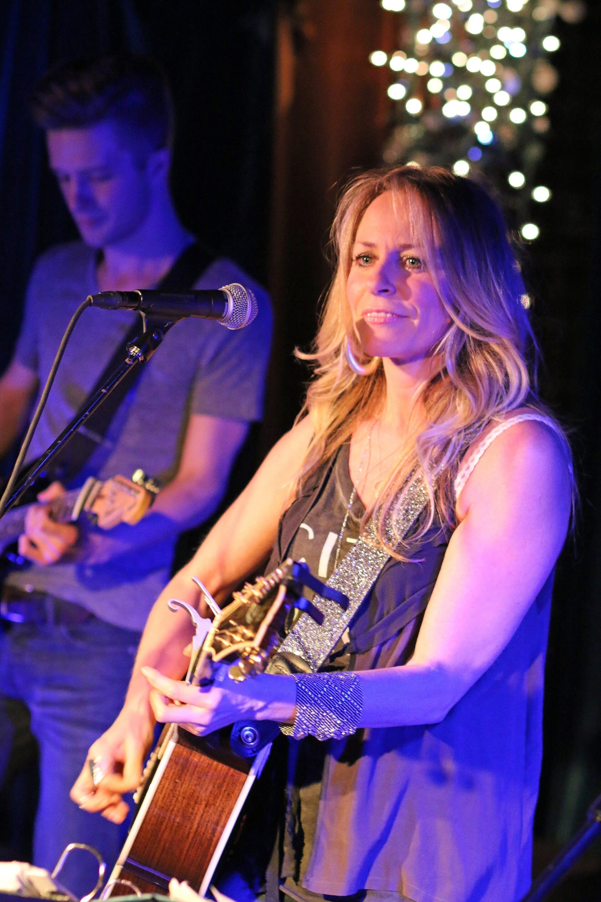 Deana Carter smiles and plays an acoustic guitar on a dimly lit stage strung with small string lights, with a bandmate playing guitar behind her.