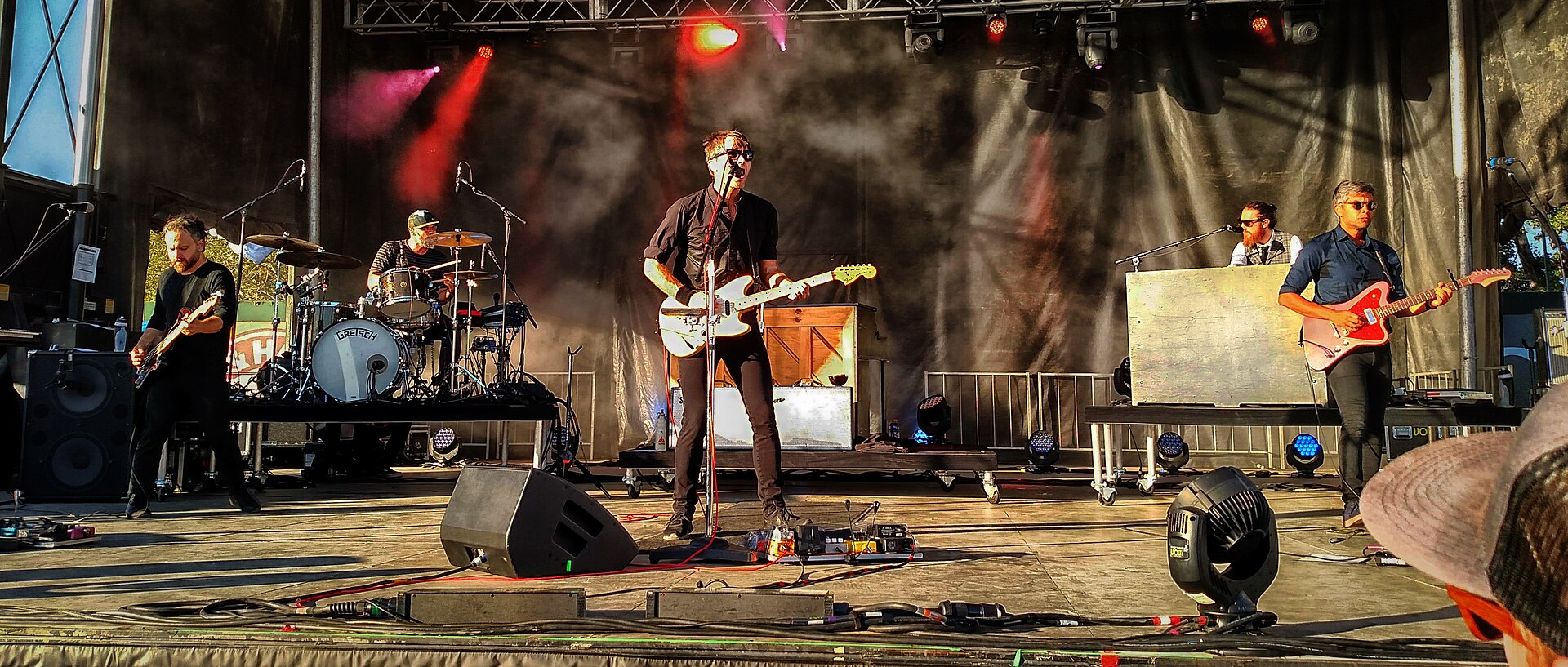 Death Cab for Cutie spread across a wide outdoor stage in late daylight, the singer at a microphone with a white Telecaster, a bassist to the left, drummer behind and two players on keys and guitar to the right.