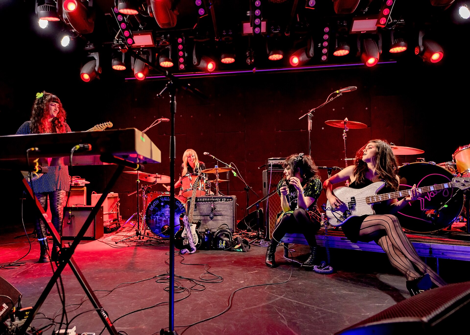 Death Valley Girls play a red-lit club stage - keyboards at left, drums behind, a singer crouched at a microphone and a bassist leaning back at right.