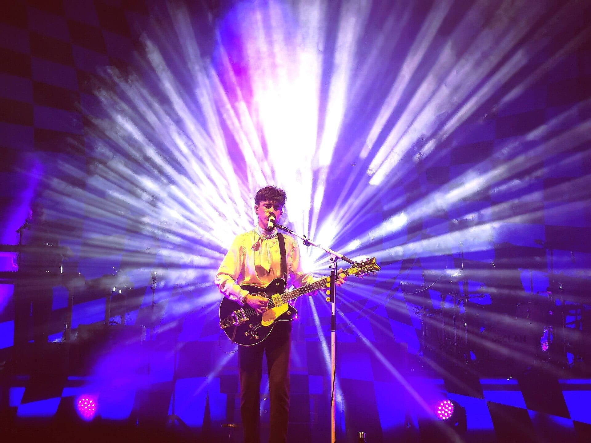 Declan McKenna sings into a microphone while playing a black electric guitar, lit by radiating purple and white stage lights.