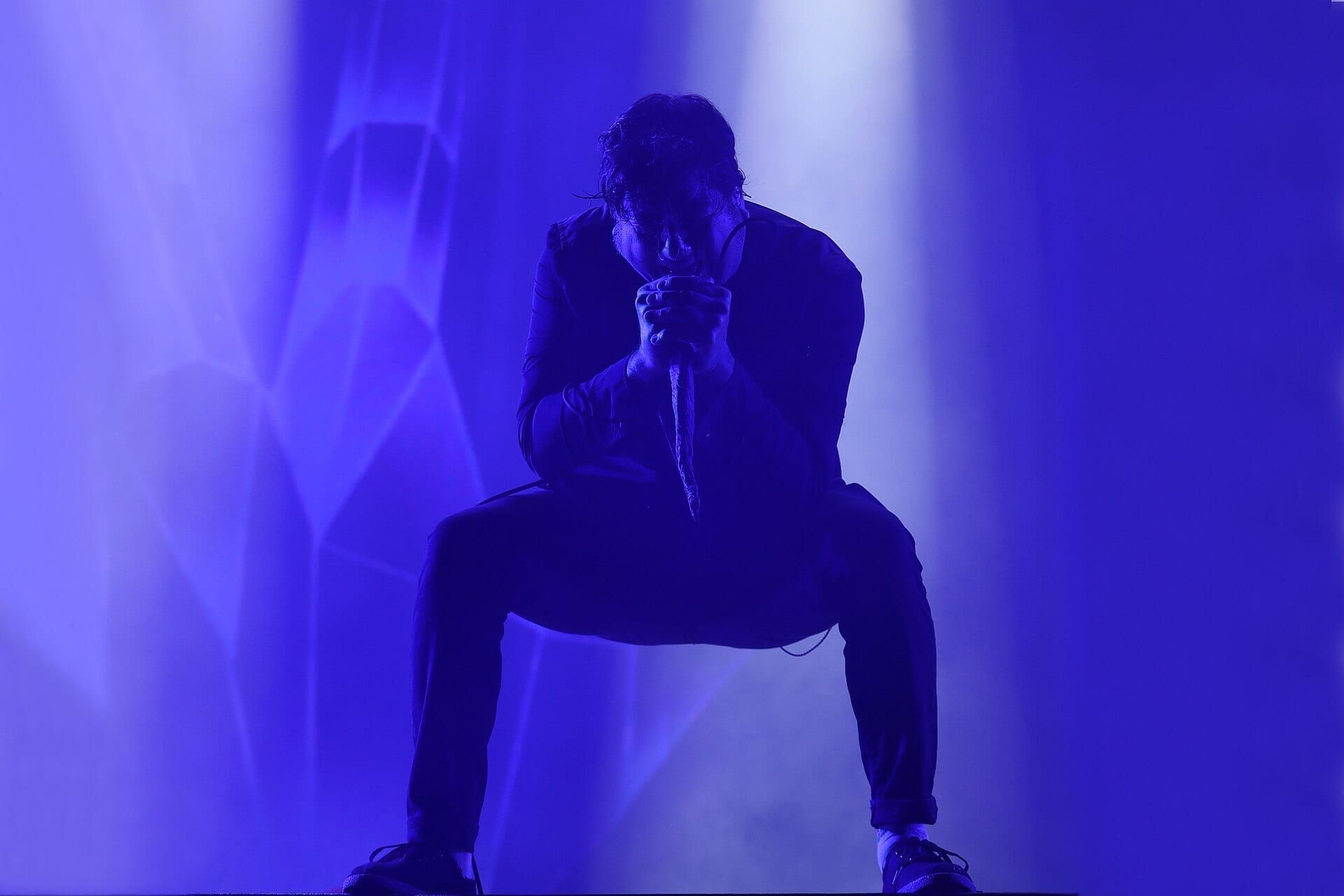 A Deftones singer crouches low over a microphone in silhouette, drenched in deep blue stage light and haze.