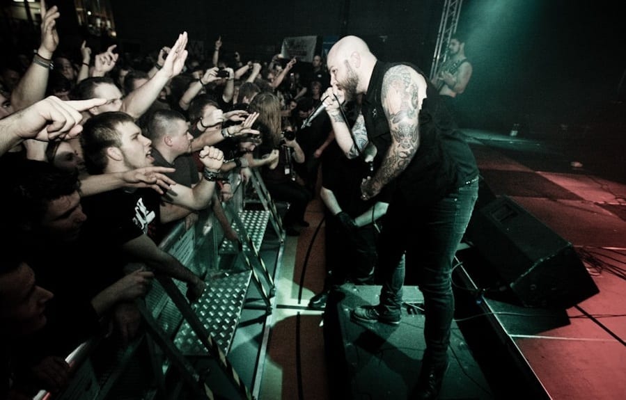 A bald, heavily tattooed singer in black leans over the barrier toward a crowd of fans with a microphone in his hand.