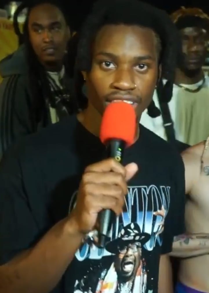 Denzel Curry, wearing a nose ring and dreadlocks, looks directly at the camera in a close-up shot.
