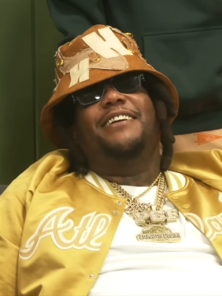 Derez De'Shon smiles while wearing sunglasses, a tan bucket hat and a yellow satin ATL jacket over gold chains.