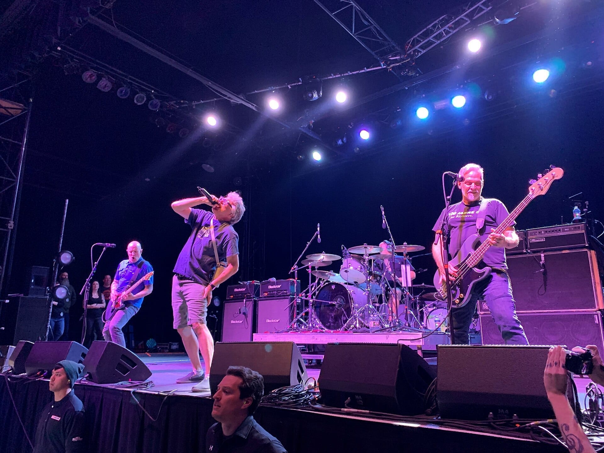 The Descendents perform on stage under purple lighting, with the singer leaning into a microphone and the bassist and guitarist flanking the drum kit.