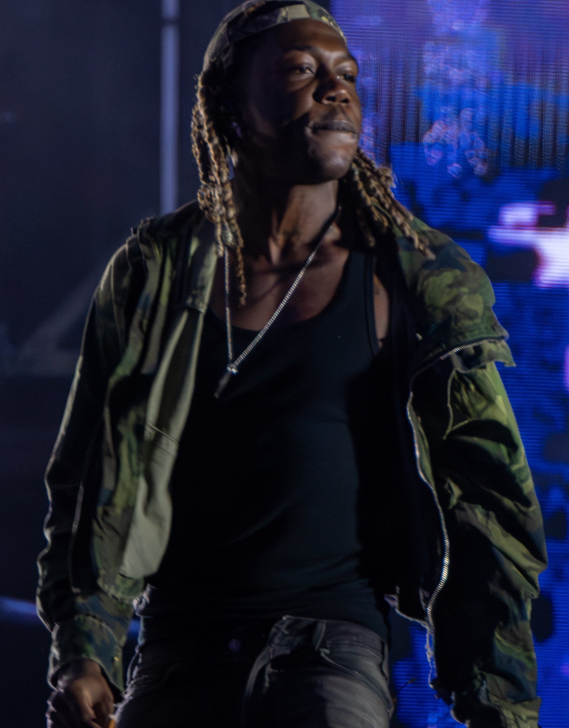 Destroy Lonely performs on stage in a camouflage jacket over a black tank top, with dyed blond braids and a chain necklace.