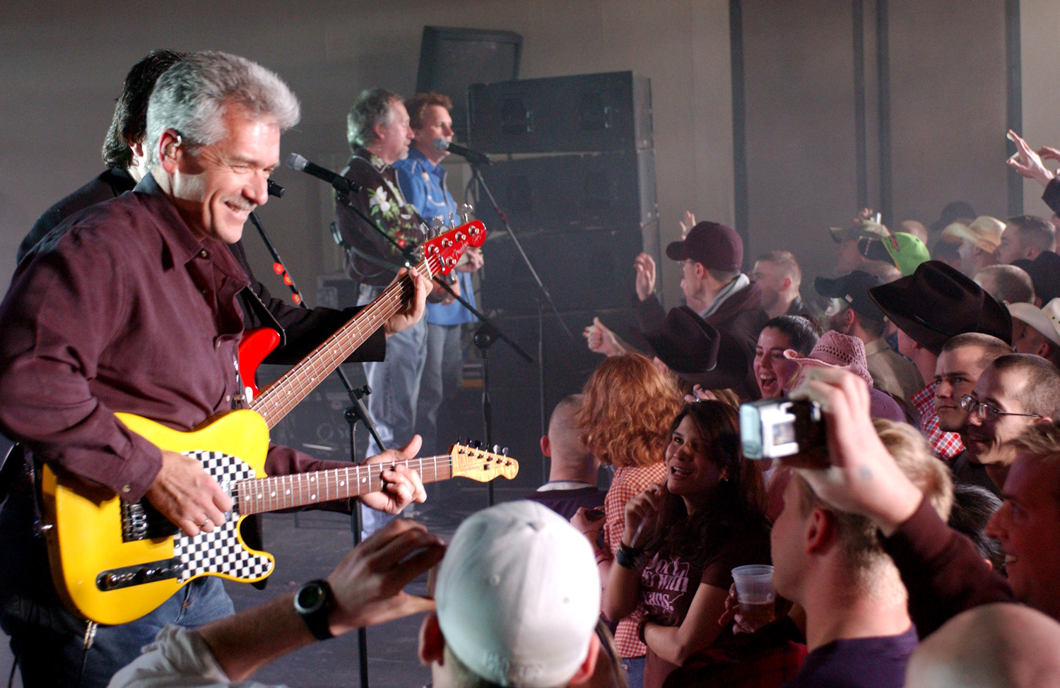 A Diamond Rio guitarist leaning toward the front of a low stage with a yellow checkered-trim Telecaster while a packed crowd of service members reaches up, two bandmates singing at microphones behind him